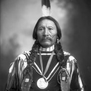 Studio portrait of Buckskin Charlie, a Native American man (Ute); dress includes fur, a hair pipe choker, a moustache, and a Rutherford Hayes Indian Peace Medal.