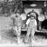 A boy and girl smile, sitting on an automobile bumper; she wears bib overalls.