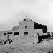 View of the Native American Laguna Pueblo Indian mission Church of San Jose, New Mexico, shows an adobe building with walled burial ground, stepped parapet, two bells and a cross.