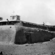 View of the mission church, Santa Ana Pueblo, New Mexico, an adobe building with vigas and twin belltowers topped with crosses.  Shows a Native American and a white man wearing a short sleeved shirt and hat.