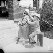 A girl sits on a stool in front of the porch of a home in Denver, Colorado. A boy is seated behind her and uses a paint brush on the back of the young girl's coat. On the ground is a palette with brushes, paint cans, and a bottle.