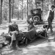 A man dressed in a sailor uniform and a woman enjoy a day outdoors as they sit on a cot in a park or forest, probably in Colorado. An automobile is parked in the background. A man walks by behind them.