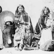 Studio portrait of Native American (Ute) women and children. Three seated women wear blanket shawls and long belted dresses, one holds a purse. Two babies are in cradleboards, two children are on the floor by the women's feet, one is sitting, the other laying down, face is hidden.