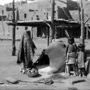 Native American (Pueblo) woman and three children stand by a beehive oven (horno), north plaza, Taos Pueblo, New Mexico. The woman wears a shawl covering her head and shoulders, there is a basket of bread by her feet. An adobe pueblo cluster is in the background.