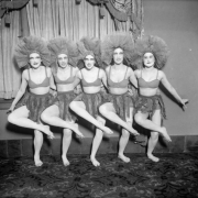 Members of the Georgia Lane Group, from the Georgia Lane School of Dancing, pose in a chorus line in Denver, Colorado; the women wear taffeta headdresses, skirts with fringe, and halter tops. Sarah Francis "Sally" Orr is in the center of the chorus line.