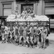 Young women and teenaged girls smile and pose in swimming suits in front of a panel truck full of snow or other material. Four of the women sit on the snow? or other material in the truck bed.