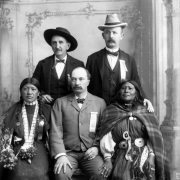 Studio portrait of two Native American (Ute) women and three white men.  The woman wearing a striped dress, shawl, concho belt, earrings, and a bead necklace is identified as Mary Tobin, mother of Mollie Cloud. The other woman wears braids, a print dress, shawl, a hair pipe choker and necklace with a toy top; she holds paper flowers in her right hand.  The men (one identified as Mr. Ward) wear suits, bow ties, felt hats, and ribbons reading: "Denver Festival of Mountain and Plain State Fair director."