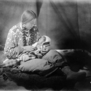 Portrait of a Native American (Crow) woman holding a baby in her lap. She sits on a buffalo hide and wears moccasins, a blanket around her waist, and a shirt decorated with shells or possibly elk teeth. The infant is wrapped in a blanket.