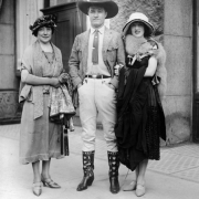 Actor Tom Mix poses with two unidentified women in Denver, Colorado. He wears a cowboy hat, boots, riding pants, coat, and tie. One woman wears a fur stole over her dress.