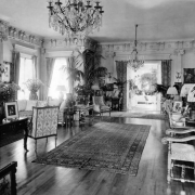 Interior view of a home, probably in Denver, Colorado. Shows crystal chandeliers, oriental rugs, potted plants, lamps, furniture, drapery, decorative columns and molding.