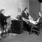 Women sit around a desk in Colorado. A woman in high heeled shoes has her feet up, by a telephone and typewriter.