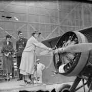 A woman christens an airplane by breaking a bottle over its propeller as men in uniform and a woman look on in possibly Denver, Colorado.
