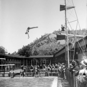 A woman dives from a tower at (probably) Eldorado Springs (Boulder County) Colorado; men in swimming suits sit by the pool.