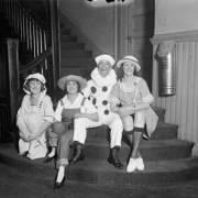 Men and women wearing costumes sit on stairs; outfits include hats, gingham, and a clown suit.