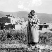 A Native American man (Taos), poses outdoors at Taos Pueblo, New Mexico. The man wears moccasins, fringed leggings, and is wrapped in a woven blanket. Adobe cluster homes and drying racks stand in the distance. The Sangre de Cristo Mountains stand in the far distance.