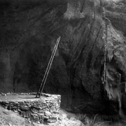 View of the entrance to a Native American (Anasazi) underground kiva in Pajarito Park (later Bandelier National Monument), New Mexico. A long wooden ladder protrudes from a square entrance to the ruins of a cylindrical stone kiva set inside a Bandelier tuff cave.  The kiva possibly stands in the side of a cliff.