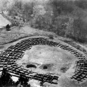 View of the Native American (Anasazi) Tyuonyi ruins in Pajarito Park (later Bandelier National Monument), New Mexico. A circular group of foundations of Pueblo style buildings circumscribe possibly three round kiva holes. A wooden rail fence separates the excavated ruins from trees and bushes growing on the adjacent mountains. A dirt road leads to the ruins.
