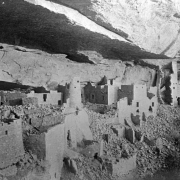 View, facing southeast, of ancient Native American (Anasazi) cliff dwellings before excavation on Chapin Mesa, Mesa Verde National Park, Montezuma County, Colorado. The stone masonry and mud mortar Cliff Palace pueblo homes stand underneath a cliffside overhang. The cylindrical and square ruins have square and T-shaped door openings. Crumbled masonry litters the ground.