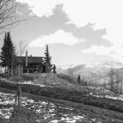 View of the Climax Mine works near Midway, Teller County, Colorado. Three men sit and stand near a wooden building with a metal roof through which a hoist protrudes. A horse stands near the building. A pile of mine tailings stands near the works. Patches of light snow cover the ground. Snow covered Pike's Peak stands in the far distance.