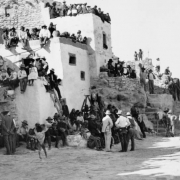 View of spectators awaiting the beginning of the Snake Dance at Walpi Pueblo, First Mesa, Arizona (Hopi Native American). Groups of men, women and children sit and stand on pueblo buildings with chimney pots as well as on adobe steps and near the dance area. Photographers and their cameras stand among the crowd.