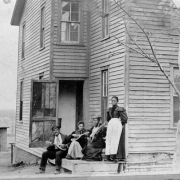 A Black man and women pose with mandolins and a guitar on the porch of a house in Denver, Colorado. A chicken and outhouse are near the two story wood frame house.