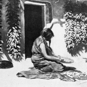 Reproduction of a painting; Native American (Hopi) woman prepares corn on a metate. Behind her is an adobe building, ristras, and pottery.