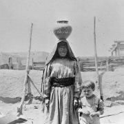 Native American (Cochiti) woman and a white boy stand in front of a pueblo wall and wire fence, Cochiti Pueblo, New Mexico. The woman carries a handcrafted pot on her head. She wears a print dress with a cloth belt and ruffled hem, and a shawl over her head. The boy wears a shirt and overalls.