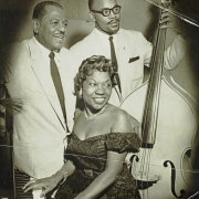 Three entertainers pose for a group photograph. Two unidentified males wear suites and ties and one plays the cello. Denver native and jazz pianist, Charlotte Mosley Cowens, sits at the piano wearing an off-the-shoulder dress and diamond jewlery.