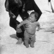 Native American (Hopi) woman leans over and stands up a large baby with her hands under his arms, Oraibi Pueblo, Arizona.