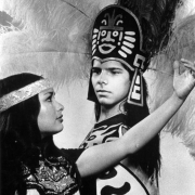 Shows a young man and woman dressed in Aztec costumes for a production presented by Fiesta Alegre de Denver and sponsored by El Centro Cultural. The man wears a feathered head piece, and carrys a shield. The woman wears a decorated bodice with fringe and a headband.