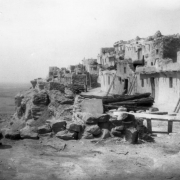 Native American (Hopi) Walpi Pueblo, Arizona with multi-story adobe and masonry structures on mesa with windows, doors, and ladders. Shows wood pile, metal bucket, ladder against a stone wall in foreground, valley in background.