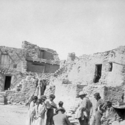 Native American (Hopi) men and children stand in a courtyard at Oraibi Pueblo, Third Mesa, Arizona. One man in the group holds a basket. The adobe and stone cluster homes have adobe stairs, wooden vigas, and wood pole cross beams supported by the walls. A tripod stands in the courtyard.