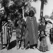A group of Native American (Navajo) men, a woman, child, and baby in a cradleboard pose outdoors near an unhitched wagon at Shiprock, New Mexico. The men are wrapped in woven blankets and wear headbands. The woman wears a dress and shawl. The girl wears a dress with a wide belt. The woman holds a baby in a cradleboard.