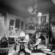 Interior view of the Drawing Room at the Palace of Governors, Santa Fe, New Mexico. Chairs, tables, a footstool, and a lamp sit in the center of the room. A boy sits in one of the chairs that line the wall. A couch sits next to the fireplace. Paintings are hung on the walls. Greenery hangs from the chandelier. Statuettes stand on the mantle.