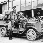 A group of Native American (Navajo) men and boys, in hats, shirts, overalls or pants,  pose on a sidewalk in Farmington, New Mexico, by the Farmington to Shiprock auto truck. The convertible truck has packages and bundles tied to the back and one man seated on a square basket; another man stands by the running board. Shows store fronts with signs that read: "Mercantile Co." and "The Rexall Store" and a large "Rexall" thermometer on wall.