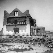 Church at Native American (Tewa) Nambe Pueblo, New Mexico; an adobe mission with double wood doors, three point facade and center belfry, balcony, attached structure and a picket fenced courtyard.