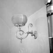 Interior view of the Hamill House, in Georgetown, Colorado. The house was built by Joseph Watson, and was acquired by William A. Hamill, his brother-in-law, in 1874. Between 1874 and 1881 Hamill made various additions to the house and grounds. The image shows a metallic light fixture attached to a wall, with a curvilinear stem with a gas regulator which supports a textured glass globe. The wall and ceiling have subtly textured wallpaper.