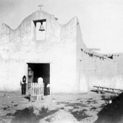 Church at Native American (Tewa) Santa Clara Pueblo, New Mexico, southwest view. Shows adobe mission with a scalloped facade and center belfry; gravemarkers in courtyard. Native American (Santa Clara Pueblo) man stands by the church entrance. He wears a shirt, pants, and vest.