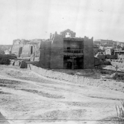 View of Mission Nuestra Senora de Guadalupe in Zuni Pueblo, New Mexico. Shows an adobe mission with a belfry and two bells, a balcony with railing over the entrance, a center support post under the balcony, vigas, and walled courtyard. Multi-story pueblo dwellings are in the distance.