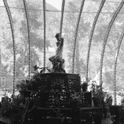 Interior view of the solarium of the Hamill House, in Georgetown,  Colorado. The house was built by Joseph Watson and acquired by mine owner William A. Hamill in 1874; shows the pyramidally tiered cast iron fernery, or plant stand, in the foreground, with potted plants on the shelves. A pewter sculpture of a nude boy blowing a horn is at the top of the stand. The decorative ribs of the solarium are behind; foliage of the trees outside shows through the glass.