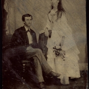 Studio portrait (sitting and standing) of a husband and wife probably on their wedding day. She holds a bouquet and wears a dress with a bustle.