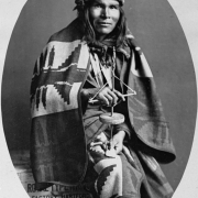 Studio portrait (sitting) of a Native American (Navajo) man. The man wears a woven blanket and holds a stone and wood hand drill. He sits on a wooden crate that reads: "[?] Factory Hartford [G]eneral Sales of [?] 253 Broadway [Ne]w York, U. S. A.".
