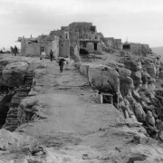 View of Walpi, Arizona, a Native American (Hopi) stone and adobe pueblo settlement; shows people, a burro, and the sandstone cliffs of the mesa.