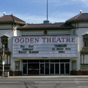 View of the Ogden Theatre (1917; Harry Edbrooke) at 935 East Colfax in the North Capitol Hill neighborhood of Denver, Colorado. Shows the two-story Mediterranean Revival-style theater.  The walls of the theater are brick and the roof is covered in red tile.  The entrance is flanked by two octagonal towers with windows, cornices, brackets, pilasters, and pediments. The marquee reads "Ogden Theatre" and "Fri Sat, Midnight, Rocky Horror," and "Tonight, La Dolce Vita, Roma."