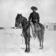 A Black Corporal, (Buffalo Soldier) in the Ninth Cavalry, poses on his horse on the Native American (Lakota Sioux) Pine Ridge Agency, South Dakota. He wears a military uniform and has a gun in a holster on his belt. Snows covers the ground and frame agency buildings show in background.