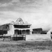 View of the Mission of San Buenaventura in Cochiti Pueblo, New Mexico, shows an adobe building with a central parapet and bell flanked by two, smaller, twin parapets, an open balcony, and an entry gate that opens to a courtyard in front of the church.