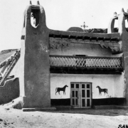 View of an adobe mission building in the Native American pueblo of San Felipe, New Mexico, shows twin bell towers, a second floor balcony, and double doors flanked by representations of horses.