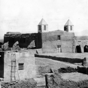 View of the Native American pueblo of Isleta, New Mexico, shows San Augustine church which features twin towers with louvered vents, adobe walls, a cross, and double doors. A ladder leans against the building; other adobe buildings nearby.