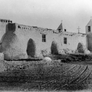 View of a mission church in the Native American pueblo of Isleta, New Mexico, shows twin bell towers with louvered vents and crosses on top, adobe wall construction, and a pile of adobe bricks.