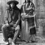 Studio portrait of an unidentified Native American Sioux man and woman in front of a painted backdrop. The man sits, wearing a three piece striped suit, neck scarf, felt hat and moccasins; the woman stands at his side wearing a striped dress and plaid shawl.